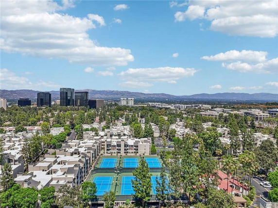 Aerial view of a residential area with multiple buildings and tennis courts, surrounded by trees and distant hills.