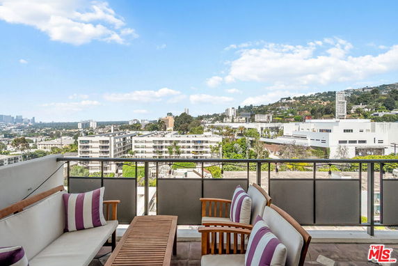 Panoramic view from a building terrace with outdoor seating, overlooking cityscape and green hills.