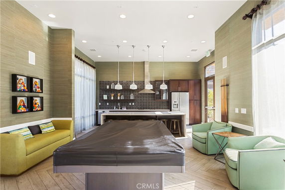 Interior view of a recreation room with a covered pool table, sofa, armchairs, and a kitchen area with bar stools.