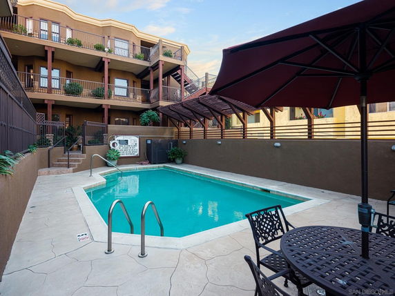 Outdoor swimming pool area with tables and chairs, surrounded by a multi-story building.
