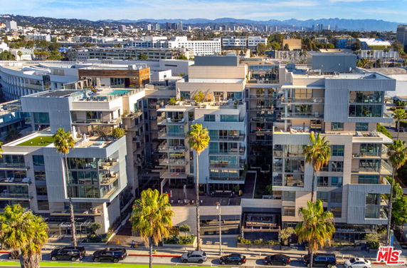 Front view of a modern multi-story apartment complex with glass balconies and palm trees lining the street.