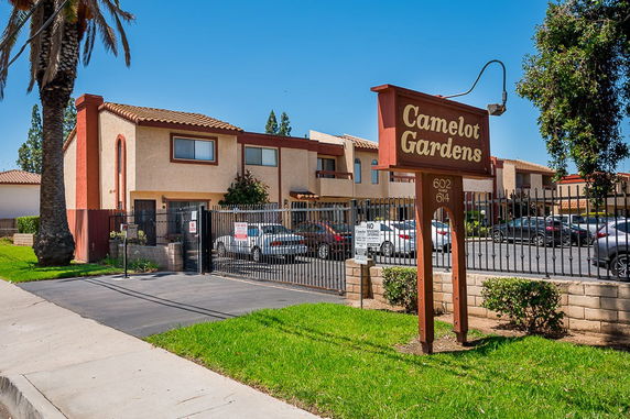 Front view of a multi-story apartment complex with a fenced entrance and a sign reading 'Camelot Gardens'.