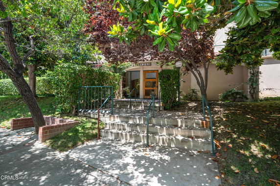 Front view of a building entrance with a staircase leading to a door, surrounded by trees and greenery.