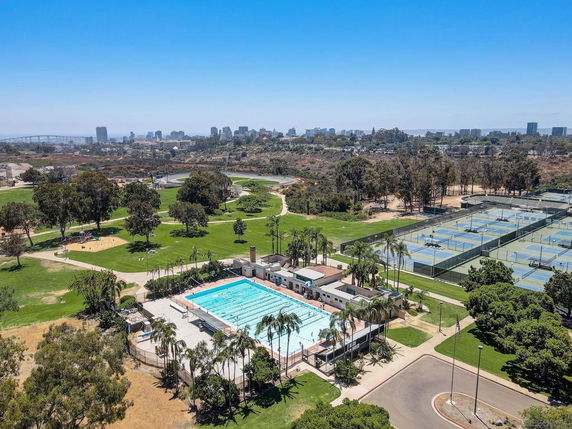 Aerial view of a public park with a swimming pool and multiple tennis courts, surrounded by green spaces and trees, with a city skyline in the background.