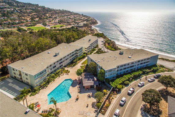 Aerial view of two large apartment buildings with a courtyard swimming pool, situated near a coastline with ocean views.