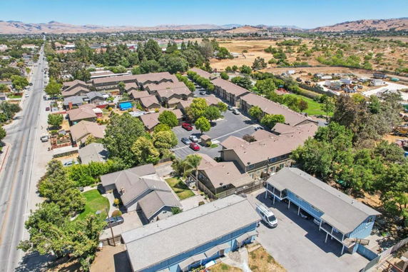 Aerial panoramic view of a residential area with multiple houses and trees, surrounded by open fields and a road.