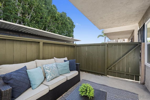Outdoor patio area with a couch, pillows, and a small plant on a rug, enclosed by a green wooden fence.