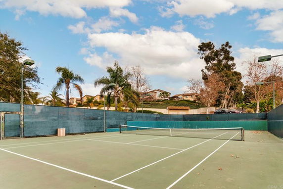 An outdoor tennis court surrounded by tall fences with palm trees and houses visible in the background.