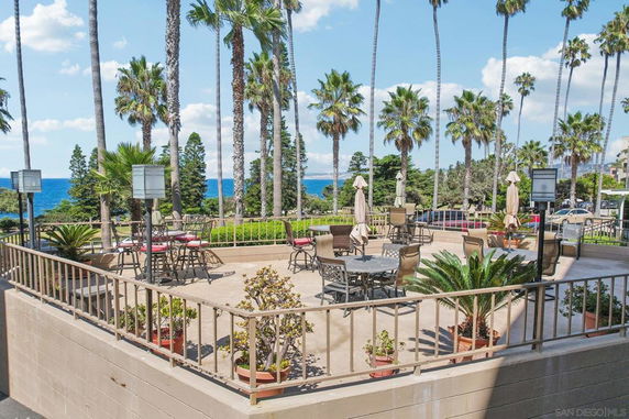 Outdoor patio area with tables and chairs, surrounded by palm trees and a view of the ocean.
