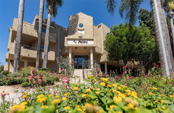 Front view of a multi-story building with balconies and a main entrance, surrounded by palm trees and plants.
