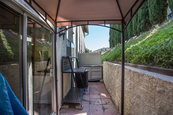 Outdoor view of a narrow patio space with a canopy, adjacent to a building wall and overlooking a hillside with greenery.