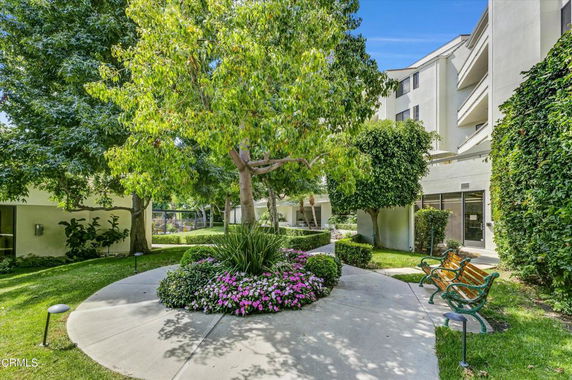 Exterior view of a multi-story building with balconies, surrounded by greenery and a landscaped garden path with benches.