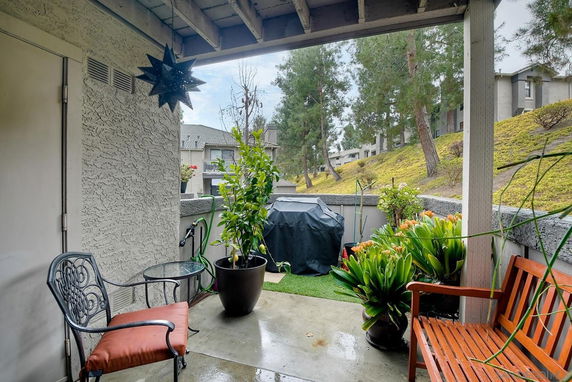 Covered outdoor patio area with benches, plants, and a grill, overlooking nearby trees and a hillside.