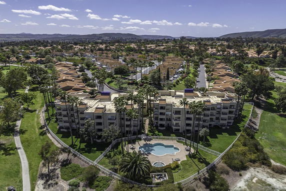 Aerial view of a residential area with a large building complex and swimming pool, surrounded by greenery and additional houses in the background.