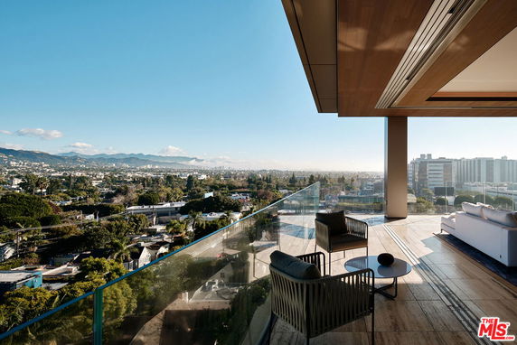 Wide angle view from a building showcasing a balcony with seating and a glass railing, overlooking a cityscape and mountains.