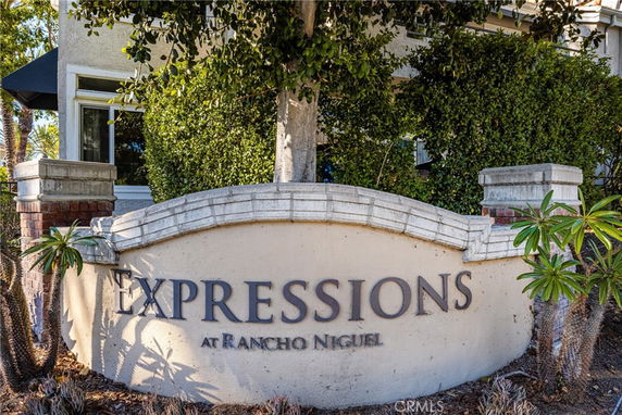 Entrance sign of a residential complex with surrounding greenery and part of a building visible.