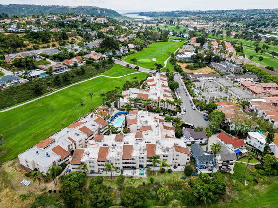 Panoramic view of a residential area with multiple buildings and a swimming pool, surrounded by green fields and hills.