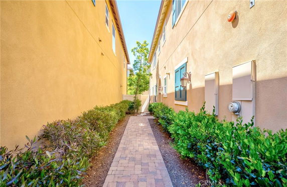 View of narrow pathway between two-story buildings with shrubs on either side.