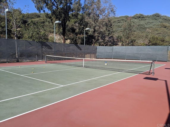Outdoor tennis court with surrounding fence and trees in the background.