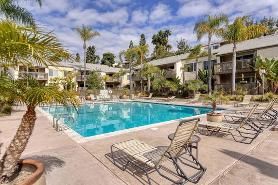 Outdoor swimming pool area surrounded by apartment buildings with lounge chairs and palm trees.