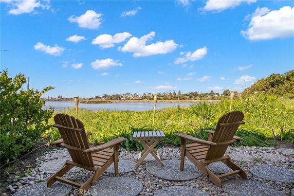 Outdoor seating area with two wooden chairs and a small table facing a water body, surrounded by greenery and under a blue sky.