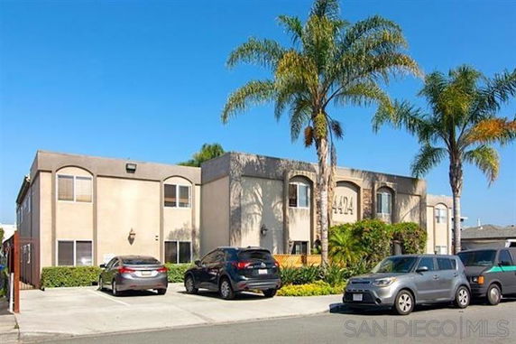 Front view of a two-story apartment building with palm trees and cars parked in front.
