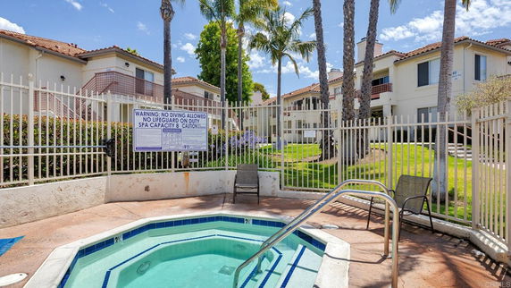 Outdoor jacuzzi area with safety sign, surrounded by a fence, and residential buildings in the background.