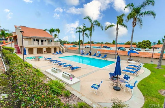 Outdoor swimming pool area with lounge chairs, umbrellas, and a multi-story building nearby.