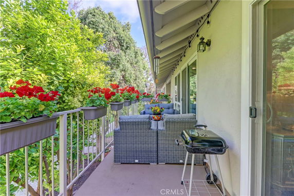 Outdoor balcony with seating, flowers, and a grill, overlooking greenery.