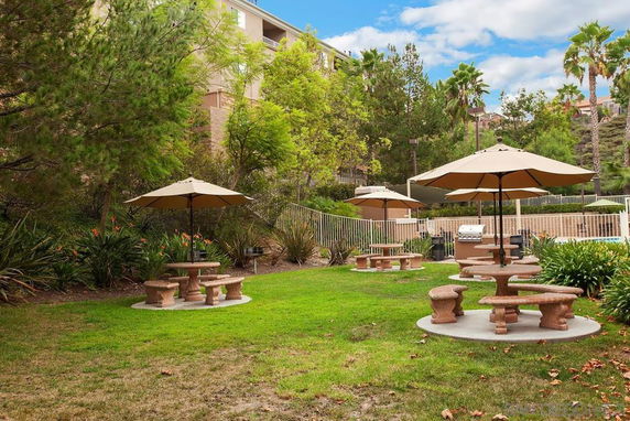 Outdoor seating area with umbrella-shaded tables and a barbecue grill, surrounded by trees and plants.