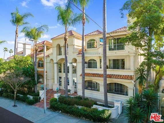 Front view of a multi-story building with balconies and arched windows, surrounded by palm trees.