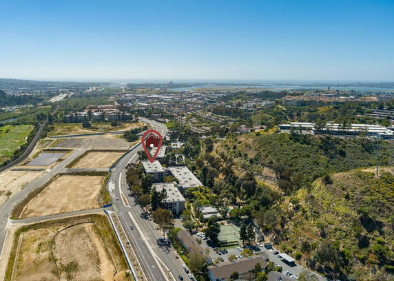 Wide angle view of a residential area with roads and buildings, surrounded by greenery and open land.