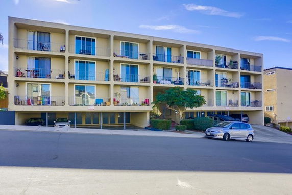 Front view of a multi-story apartment building with balconies and parking area in front.