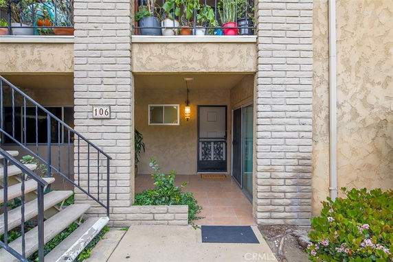 Front view of a residential entrance with stairs and potted plants on the balcony above.