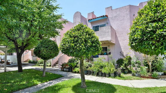 Front view of a two-story house with pink walls and a small balcony, surrounded by green plants and trees.