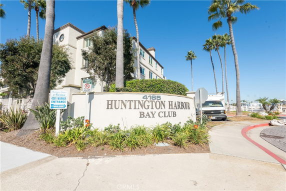 Front view of a multi-story building with palm trees and a sign that reads '4165 Huntington Harbour Bay Club.'