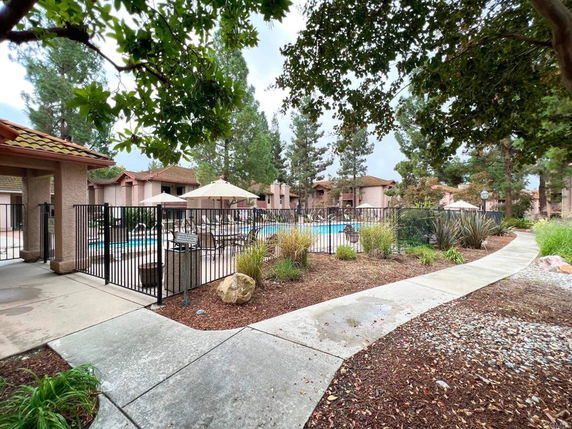 Outdoor swimming pool area surrounded by a fence with nearby residential buildings and tall trees.