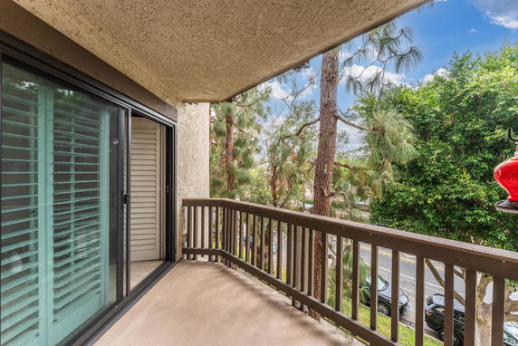 Balcony view from an apartment showing sliding glass doors with shutters, overlooking trees and a street.
