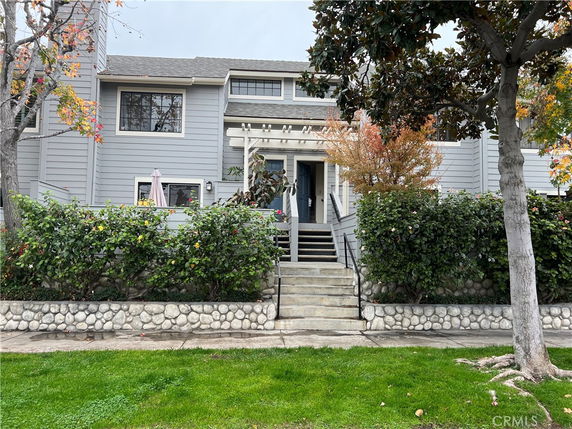 Front view of a two-story house with gray siding and a stone base. The entrance is up a set of stairs with a wooden railing.
