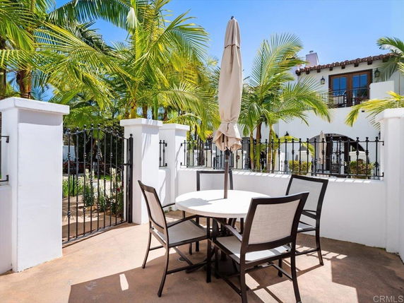 View of a white house with a balcony and palm trees in the surroundings. A patio set with a table and chairs is in the foreground.