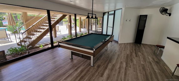 Interior view of a room with a billiards table, wooden flooring, and large windows leading to an outdoor area.