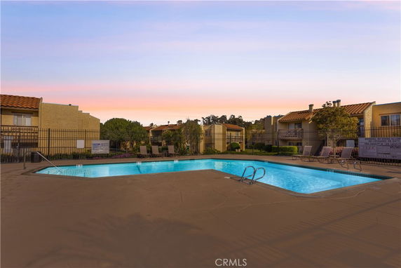 Outdoor swimming pool area surrounded by buildings with lounge chairs and a fence.