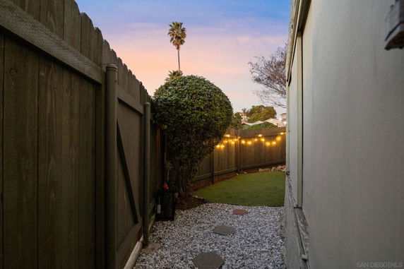 Narrow outdoor area with fencing, a gravel path, and string lights.