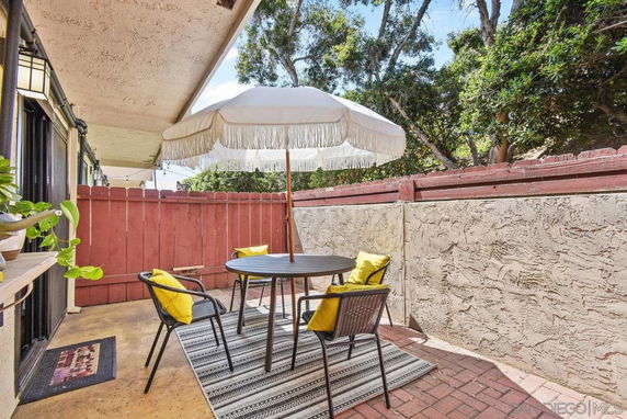 Outdoor patio area with a circular table, chairs, and an umbrella, surrounded by a wooden fence and trees.