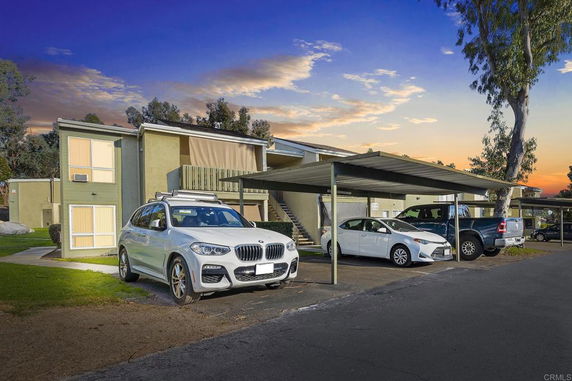 Outdoor parking area with several cars parked under a shade next to a residential building.