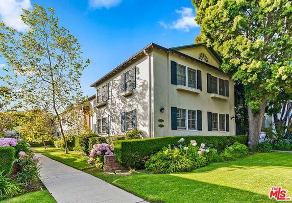 Front view of a two-story house with green shutters and surrounded by shrubs and trees.
