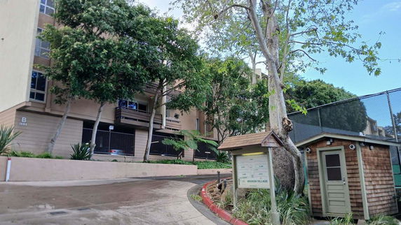 Front view of a multi-story residential building with trees and a small wooden structure nearby.