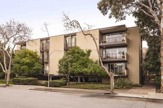 Front view of a multi-story apartment building with balconies surrounded by trees.