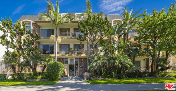 Front view of a multi-story residential building with balconies and tall trees in front.