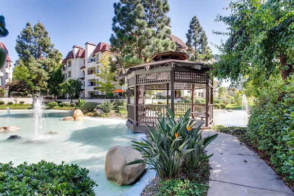 Outdoor view with a water fountain, gazebo, trees, and multi-story buildings in the background.